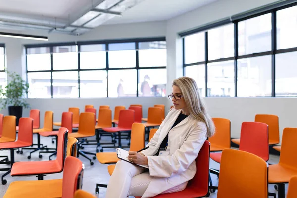 一个有着一头金发的高加索女医生 穿着漂亮的衣服和眼镜 坐在一间空荡荡的现代会议室的椅子上 在一张纸上写字的侧视图 — 图库照片