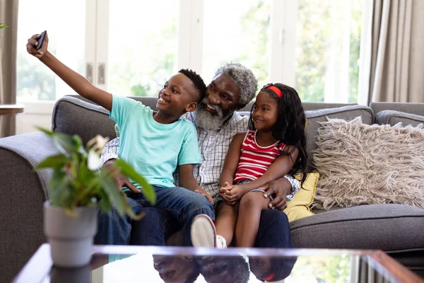 上了年纪的非裔美国人和他的孙子孙女们一起享受他们在家里的时光 坐在沙发上 用智能手机 自私自利 — 图库照片