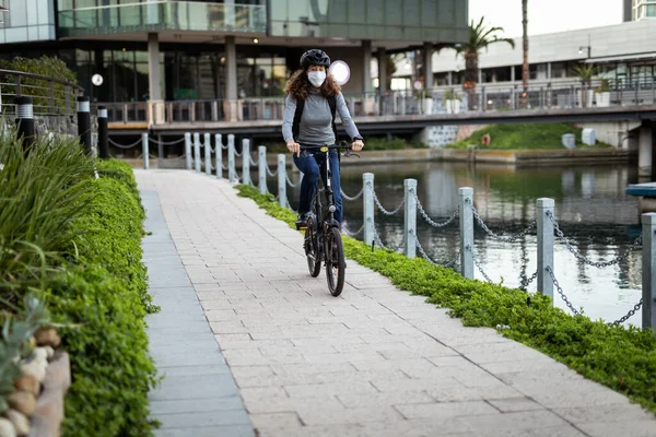 Caucasian woman out and about in the city streets during the day, wearing a cycling helmet and a face mask against covid19 coronavirus, cycling beside a canal