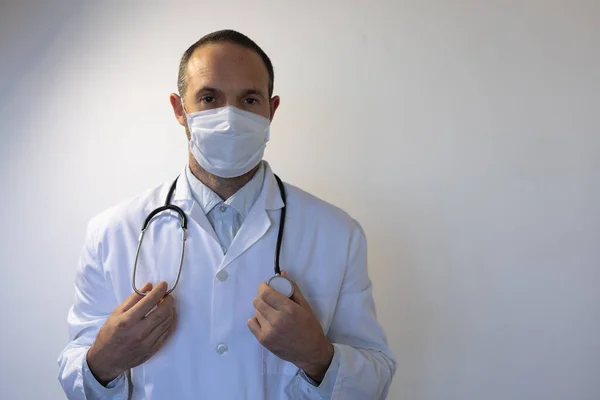 医者の制服を着た白人男性の肖像画と肩の聴診器 顔のマスク カメラを見て空の白い部屋に立って — ストック写真