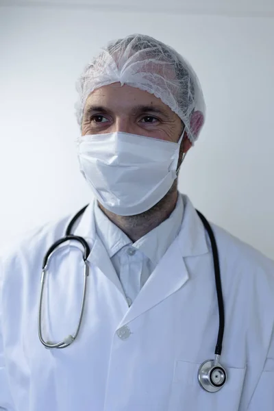 身穿医生制服 肩上戴听诊器 戴着面具 站在一间空荡荡的白色房间里的白人男子 — 图库照片