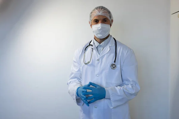 肩に医者の制服と聴診器を着て顔のマスクをして白い部屋に立って手袋をしている白人男性の肖像画 — ストック写真