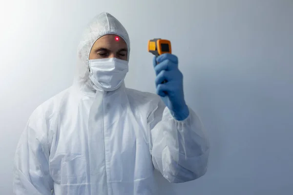 白种人穿着实验室工作服 戴着面具 站在一个空荡荡的白色房间里 戴着手套 量着体温 — 图库照片