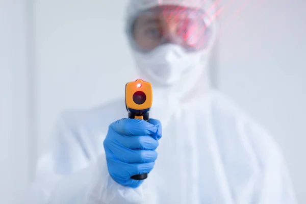 一名身穿防护服 面罩和手套的白人女性保健工作者的画像 她戴着一只非接触式温度计 直视着摄像机 — 图库照片
