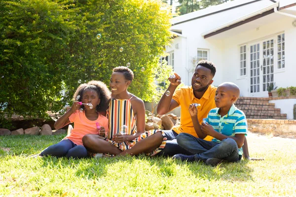 非裔美国男人和女人花时间与他们的儿子和女儿在他们的花园 坐在草坪上 制造肥皂泡 隔离隔离中的社会疏离和自我隔离 — 图库照片