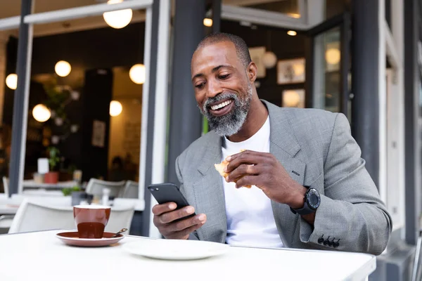 アフリカ系アメリカ人の男性がカフェでコーヒーテラスに座って軽食をとりスマートフォンを使って — ストック写真