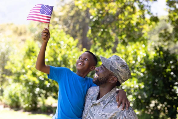 在阳光灿烂的一天 一名身穿军装 抱着儿子 抱着儿子 看着相机 面带微笑的非裔美国男子举着一面迷你国旗 — 图库照片