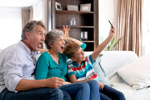 老白种人夫妇和他们的孙子呆在家里 坐在客厅的沙发上 拿着智能手机自拍 — 图库照片