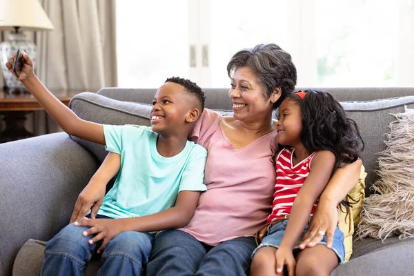 高齢の混合人種の女性と彼女の孫は一緒に自宅で彼らの時間を楽しんで ソファに座って 笑顔を受け入れ スマートフォンを使用して 自撮りを取ります — ストック写真