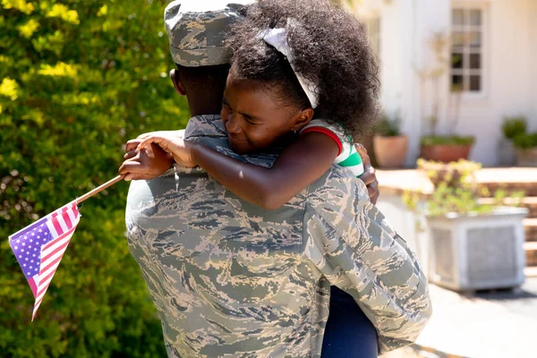 在阳光灿烂的一天 身穿制服 手持美国国旗拥抱女儿的美国黑人士兵站在房子旁边 — 图库照片