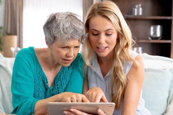 白种人的老妇人穿着绿色的衬衫 她的成年女儿留着一头长长的金发 一起呆在家里 坐在客厅的沙发上 用平板电脑 — 图库照片