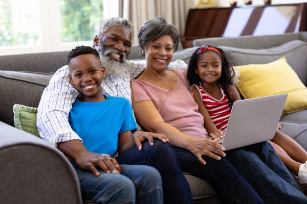 上了年纪的混血种族夫妇和他们的孙子孙女在一起 享受他们在家里的时光 坐在沙发上 使用笔记本电脑 — 图库照片