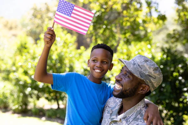 アフリカ系アメリカ人の男は軍の制服を着て帰国し息子を抱きかかえてカメラを見て笑っている男の子は晴れた日にミニフラグを持っている — ストック写真