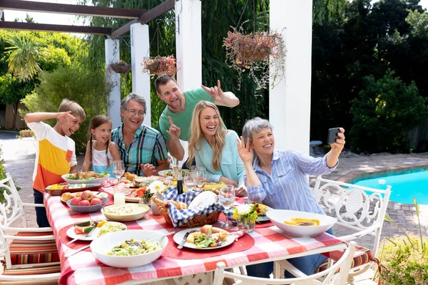 在阳光明媚的日子里 白种人三代一家人在花园里共度时光 坐在餐桌旁吃午饭 而一位老妇人则拿着智能手机在自作主张 — 图库照片