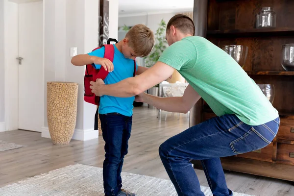 一名身穿绿色T恤的白人男子帮助他的儿子穿着红色背包准备上学 — 图库照片