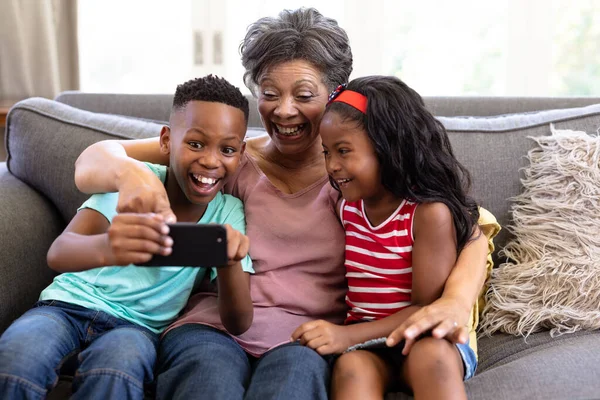 上了年纪的混血种族女人和她的孙子孙女们一起享受他们在家里的时光 坐在沙发上 用智能手机 自私自利 — 图库照片