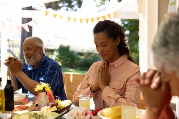 目を閉じて祈りの手でテーブルに座っている多民族 多世代の家族の側面ビュー 太陽の下でパティオの外で一緒に食事をする前に恵みを言います — ストック写真