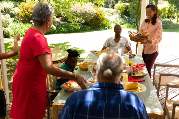 多民族 多世代の家族が食べ物を提供し 一緒に太陽の下でパティオの外で食事のためのテーブルに座っているの側面ビュー 祖母と母親が立ってテーブルに食べ物を持って来て 他の人は座って — ストック写真