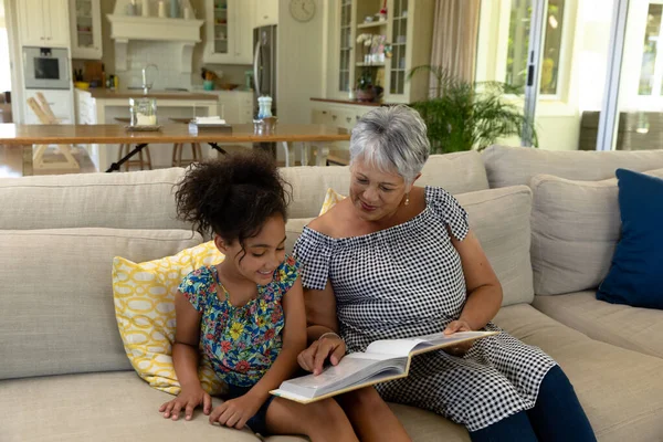 彼女の若い孫娘とソファの上に座って本を持って一緒にそれを読んでいます — ストック写真