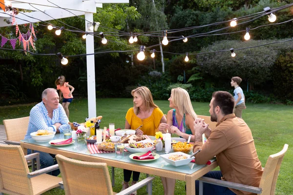 庭の白人の多世代の家族の側面図 大人が一緒に食事のためにテーブルに座って話をしながら 孫娘と孫が背景で遊ぶ — ストック写真