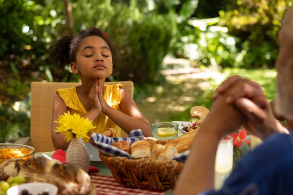 目を閉じて祈りの手でテーブルに座っているアフリカ系アメリカ人の少女の正面図は 太陽の下でパティオの外で家族と一緒に食事をする前に 恵みを言って フォアグラウンドで彼女の祖父 — ストック写真