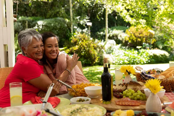 太陽の下でパティオの外の家族の食事のためのテーブルに座って抱擁し 笑顔の混合レースの女性と彼女のシニアの母親のフロントビュー — ストック写真