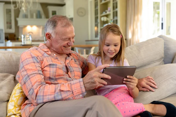 一个白人老人和他的孙女坐在客厅的沙发上 一起使用平板电脑 面带微笑 — 图库照片