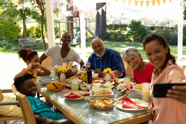 太陽の下でパティオの外で一緒に食事をするテーブルに座っている多民族 多世代の家族のフロントビュー 彼らと一緒にすべての笑顔で自撮りを取るためにスマートフォンを使用して混合レースの母親 — ストック写真