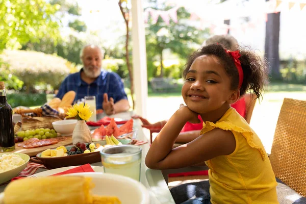 太陽の下でパティオの外の家族の食事中にテーブルに座っているカメラに向かって黄色のドレスの回転と笑顔を身に着けている若い混合レースの女の子の側面図 彼女の祖父母は背景に座って — ストック写真