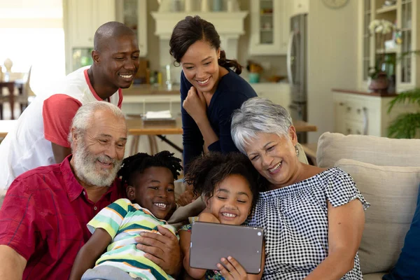 孫が持っているタブレットコンピュータを見てソファの上に集まったリビングルームの自宅で多世代の混合レース家族のフロントビュー — ストック写真