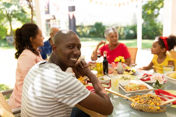 アフリカ系アメリカ人の男性がカメラを向けると笑顔を浮かべ 太陽の下でパティオの外で多民族多世代の家族と食事中にテーブルに座って — ストック写真