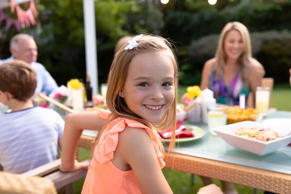 一个白人女孩坐在桌旁 和她的多代家人在花园里吃饭时对着相机微笑的画像 — 图库照片