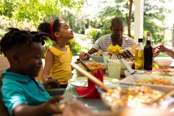 一个多族 多代同堂的家庭 双目紧闭 手牵手坐在餐桌旁 在阳光下与一位年轻的非裔美国男孩手牵着手 在外面的庭院里一起进餐之前 一边做着祈祷 — 图库照片