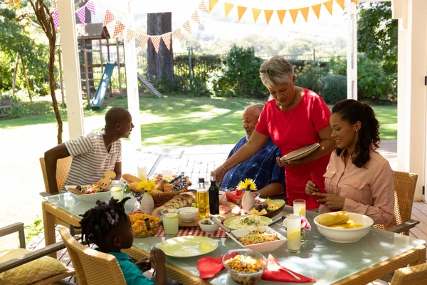 多民族の多世代の家族が食べ物を提供し 太陽の下でパティオの外で一緒に食事のためにテーブルに座っているの側面図 祖母は立ってテーブルの上にプレートを置く — ストック写真