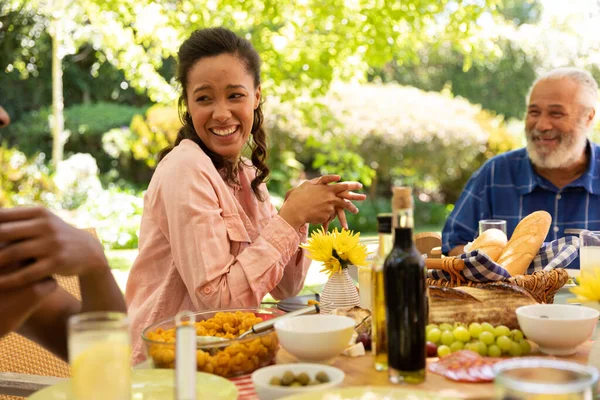 太陽の下でパティオの外で家族の食事中にテーブルに座って笑顔混合レースの女性と彼女のシニア父親の側面図 — ストック写真