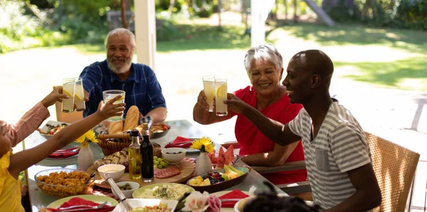 太陽の下でパティオの外で一緒に食事をするためにテーブルに座っている多民族 多世代の家族の側面図 レモネードのグラスを上げ トーストを作る — ストック写真