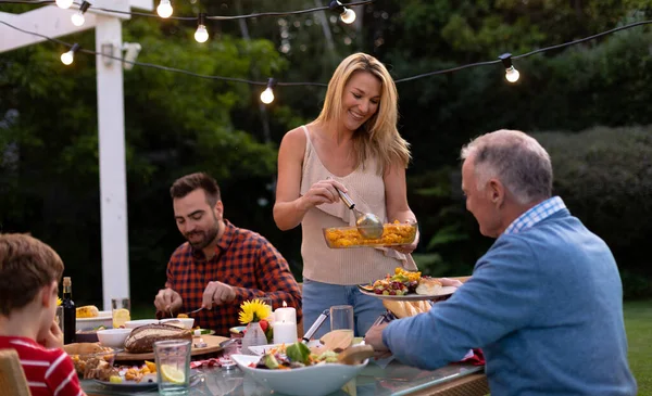 夕食のテーブルで外の多世代の白人の家族の前で食事をしたり 座ったり 食事をしたり 食事をしたり お互いに料理を提供したり 母親は立っていると彼女のシニア父親にサービスを提供しています — ストック写真