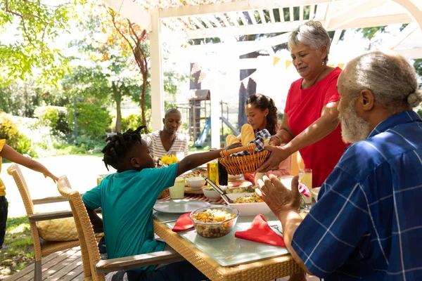 多民族 多世代の家族が食べ物を提供し 太陽の下でパティオの外で一緒に食事のためにテーブルに座って 祖母は立って 彼女の孫にいくつかのパンを提供 — ストック写真