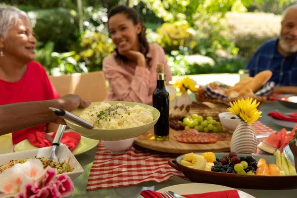 テーブルに座って食事を提供する多民族の大人の家族の側面ビュー 太陽の下でパティオの外で一緒に食事中に話して笑顔 — ストック写真