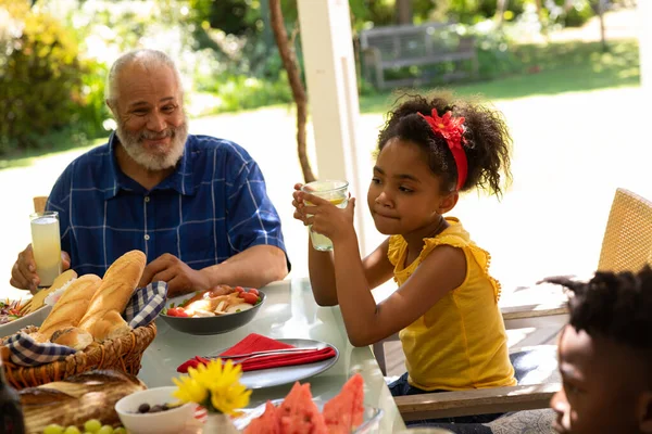 弟と笑顔の祖父とテーブルに座っているガラスを持っている若いアフリカ系アメリカ人の少女の側面図太陽の下でパティオの外で家族の食事のために — ストック写真