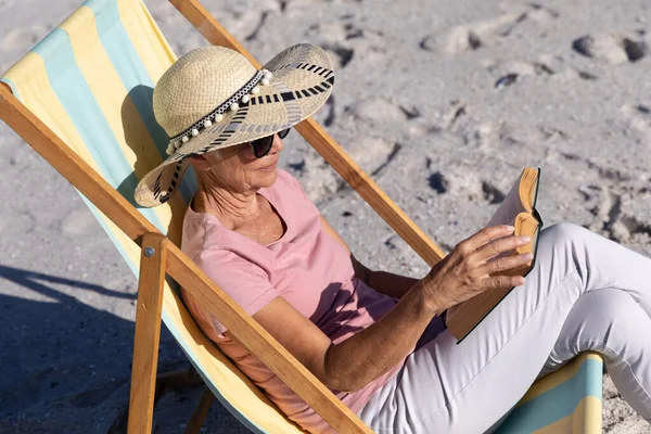 年长的白人妇女在海滩上享受时光 坐在甲板上的椅子上看书 — 图库照片