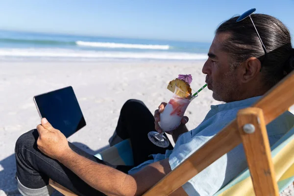 年长的白人男子在海滩上享受时光 坐在甲板上的椅子上 使用数码平板电脑 — 图库照片