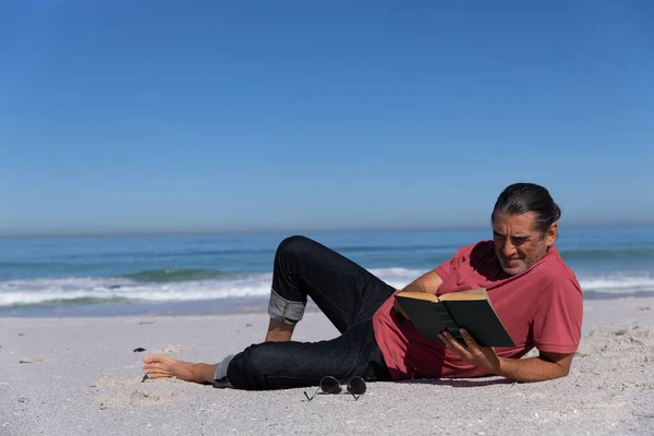 Homem Caucasiano Sênior Aproveitando Tempo Praia Deitado Areia Lendo Livro — Fotografia de Stock