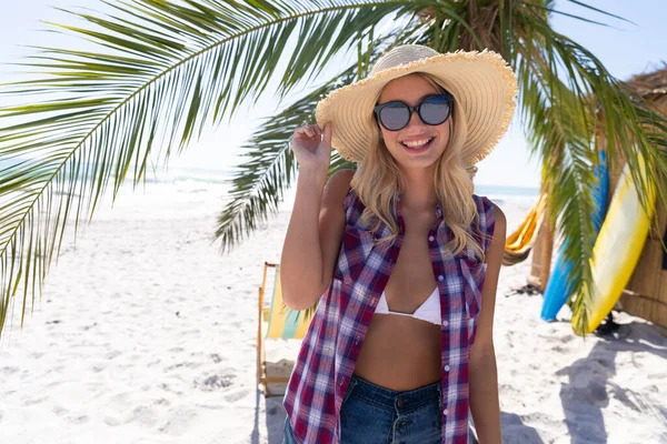 白人妇女在海滩上享受时光 站在棕榈树旁 拿着帽子 面带微笑 — 图库照片