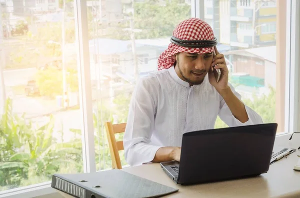 young Arabian business man talking on mobile phone and working finance about cost with laptop computer and document on desk at home office, financial, business technology and investment concept