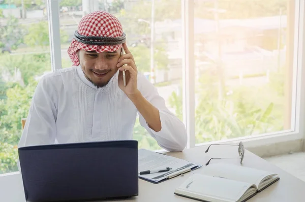 young Arabian business man talking on mobile phone and working finance about cost with laptop computer and document on desk at home office, financial, business technology and investment concept