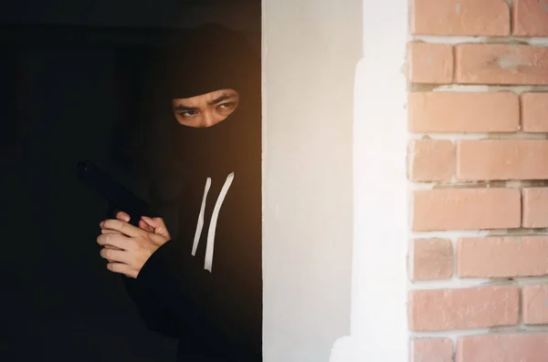 thief in black jacket and mask hood with gun hiding in corner of building waiting and looking for victim, violence, crime, robber and steal concept