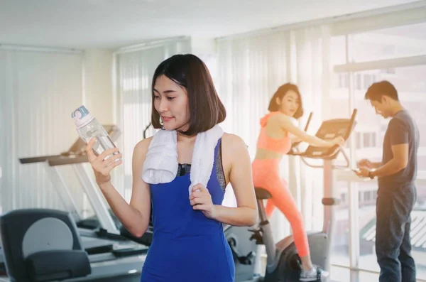 young happy smile asian woman slim body with towel drinking water while resting after workout for good healthy in fitness gym, bodybuilder, healthy lifestyle, exercise and sport training concept