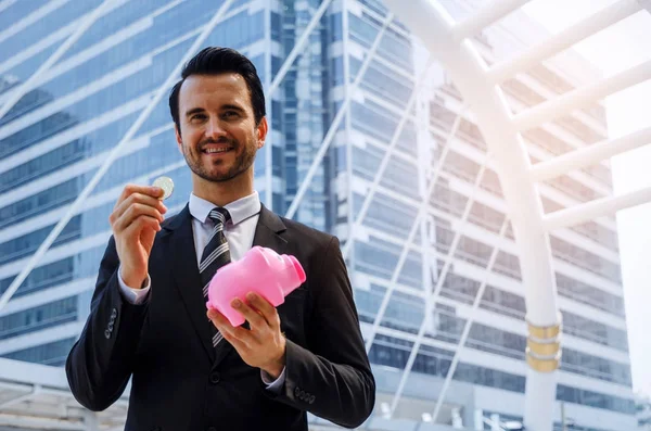 caucasian smart business man in black suit holding piggy bank and golden Bitcoin on modern city background, digital technology, virtual currency, financial, investment, payment, saving money concept