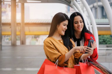 iki güzel neşeli Asya ve beyaz genç kadınla mobil akıllı telefon ve birlikte modern şehir, kış indirimi, Cuma gecesi, yürüyüş poşetler ödeme ve alışveriş online kavramı holding